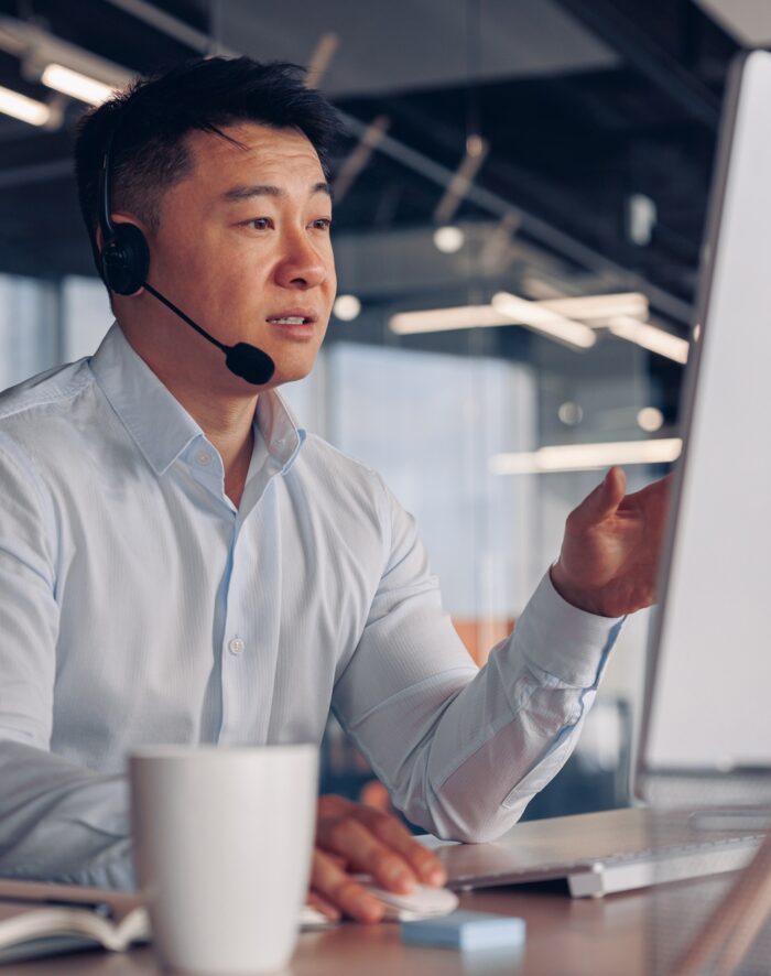 Focused asian businessman in headphones talking online with client sitting in office