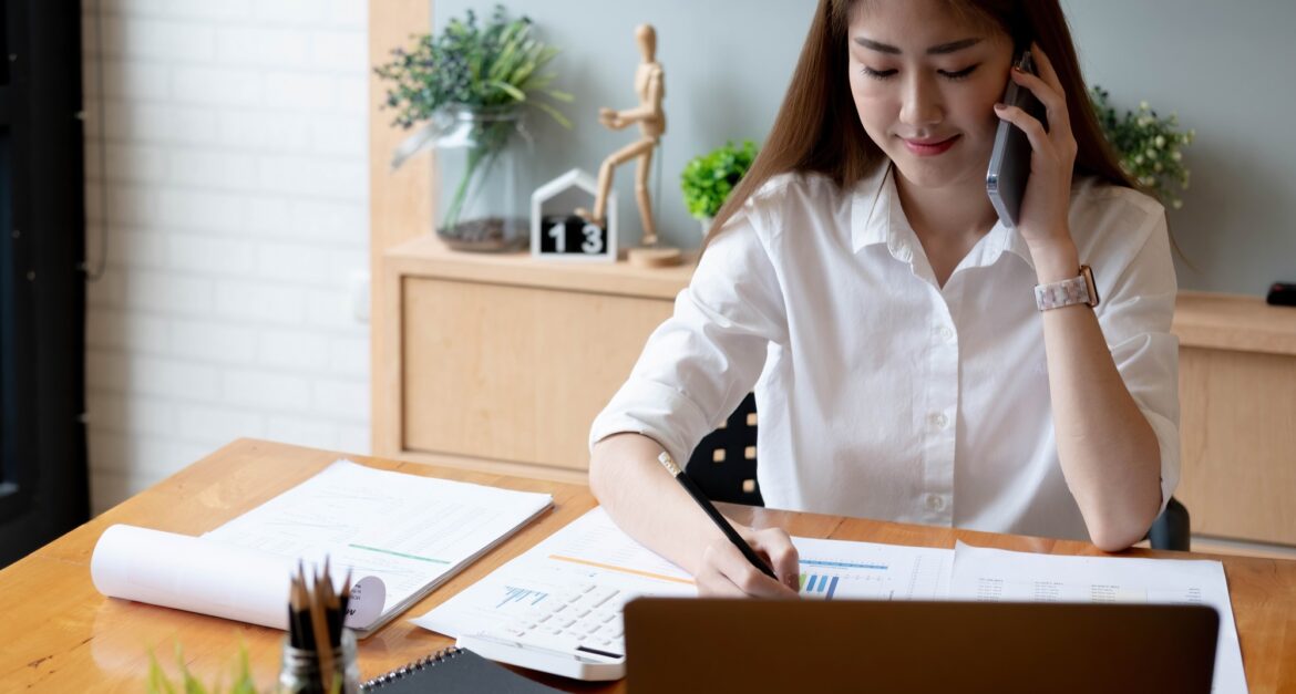 Smiling asian woman taking call on mobile phone during working on laptop computer for financial