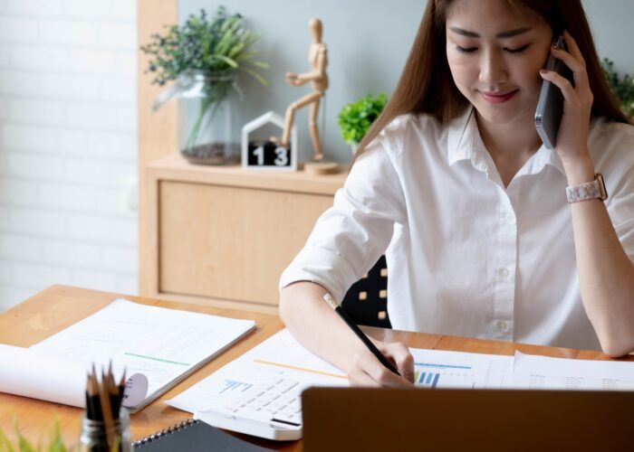 Smiling asian woman taking call on mobile phone during working on laptop computer for financial